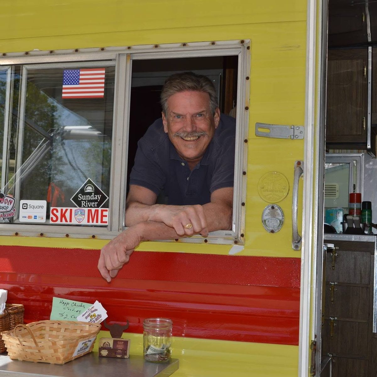 Ron Demers leaning out the service window of his yellow and red 1973 camper at Rocket Ron's American Grill in Oxford, Maine