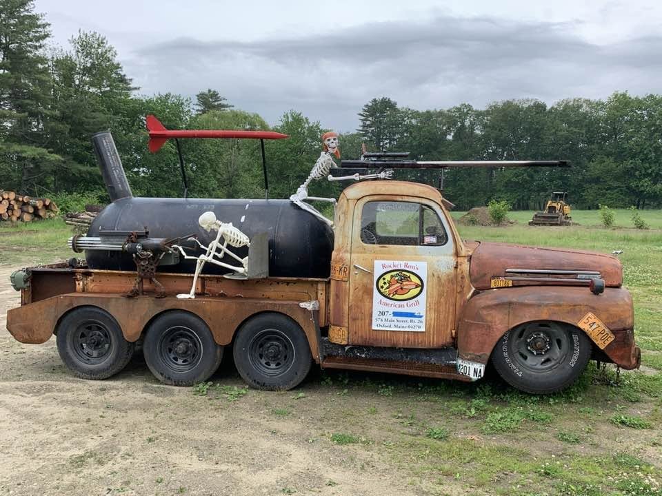 Rocket Ron's rat rod smoker truck: a rusted flatbed carrying a barrel smoker, a red rocket fin, and two skeletons riding along